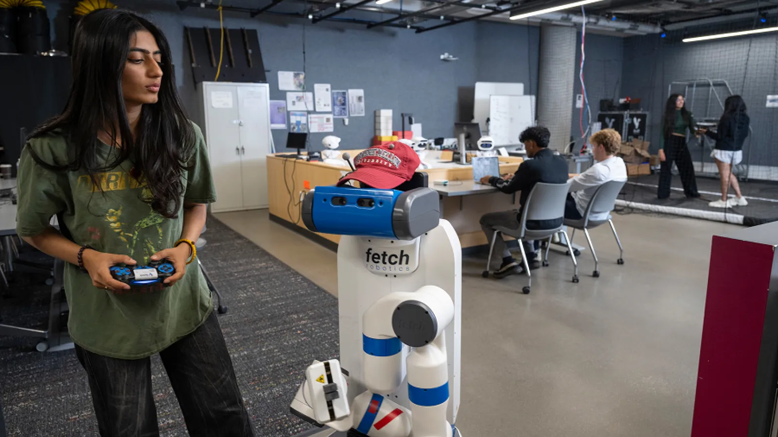 Woman holding a controller beside a humanoid robot in a modern robotics lab; coworkers sit at desks behind her, creating a collaborative, futuristic mood.
