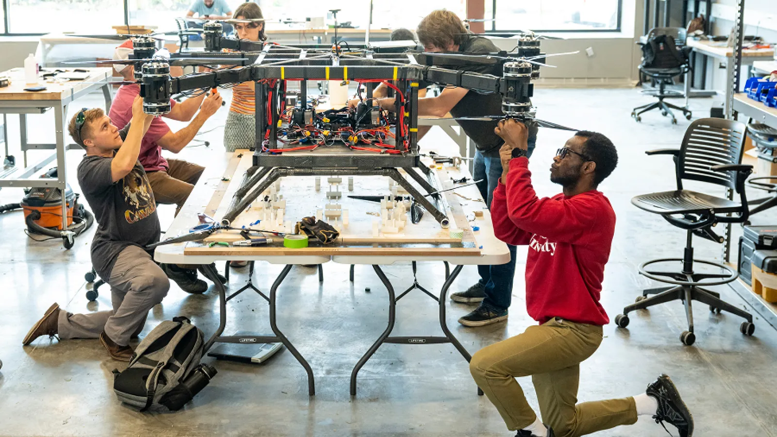 Engineering students collaborate in a workshop, assembling and testing a large custom drone with exposed wiring and quadcopter arms.