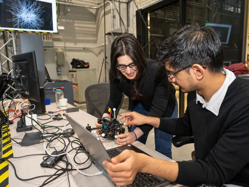 Two engineers collaborate on a small robot prototype at a workshop desk, surrounded by cables and monitors, focused and engaged.