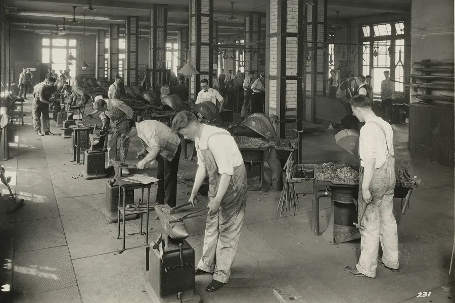 Students focus over metal anvils in a large workshop.